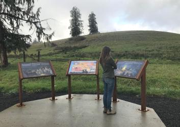 A person with long hair and wearing a T shirt and jeans looks at one of three interpretive panels set in a semi-circle on a concrete pad. A grassy hill with a few sparse evergreen trees on it rises under a cloudy sky in the background.