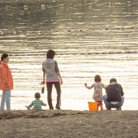 Family with 2 young children and 3 adults exploring the sand at Sunset Beach. One adult is wearing light colored jeans and a peach colored jacket, another is in black track pants and a grey and pink sweatshirt, and the third in a grey or blue sweatshirt and jeans. One of the youth is wearing a green and white stripped shirt and the other is in a floral pattern. The swimming buoys are visible on the water and it appears to be golden hour where the sun creates a soft, golden glow. 