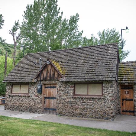 Restroom building with rock walls and wooden doors with heavy black hardware, a wood shake roof with moss growing in spots. Green trees stand tall behind the building and a cloudy sky