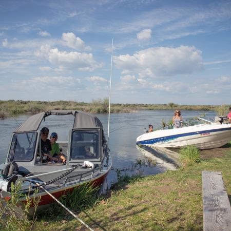 Two boats beached on a grassy beach with a picnic table in view. Two fishermen are smiling in one boat and the other boat is being pushed into the water by two men while two women wait in the boat.