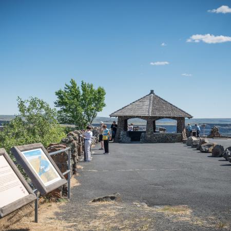 Looking at the rock shelter with a shake roof, with trees and interpretive panels lining the walkway. A blue sky set in the background. 