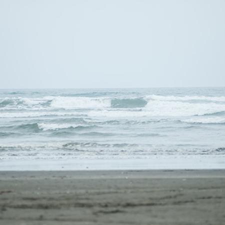 A flat, sandy beach meeting with rolling Pacific Ocean waves under a pale blue sky