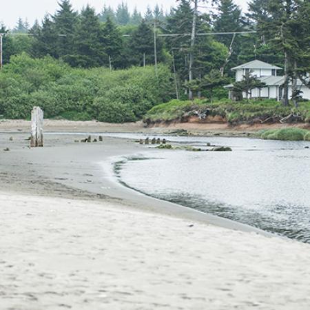 A river flowing through a sandy shoreline toward the ocean. Trees, and seaside houses are in the distance