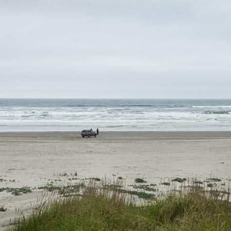 A wide, flat sandy shoreline framed above with low, rolling ocean waves and below with shrubby dune grass