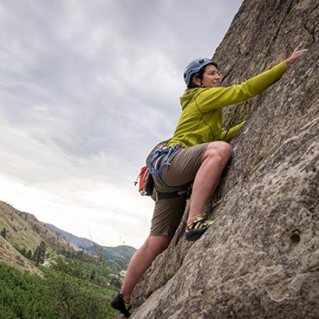 A person in a yellow shirt and shorts and wearing a helmet scales a granite rock face. A hillside rises in the distance under a cloudy gray sky.