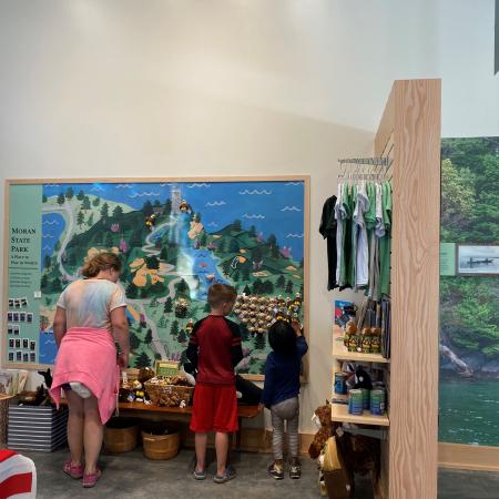 A girl wearing a tie dyed shirt with a pink sweater wrapped around her waist and pink shoes, a boy wearing a red shirt with blue sleeves, red shorts and blue slid on shoes, and a little girl wearing a long blue shirt, black and white stripped pants with blue sneakers look at small stuff animals on a shelf and hung on the wall.