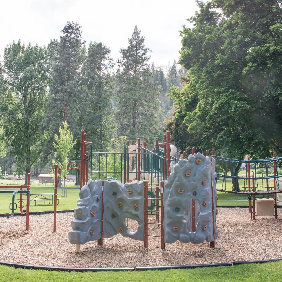 A children's playground with a large lawn, picnic tables, and trees in the background.