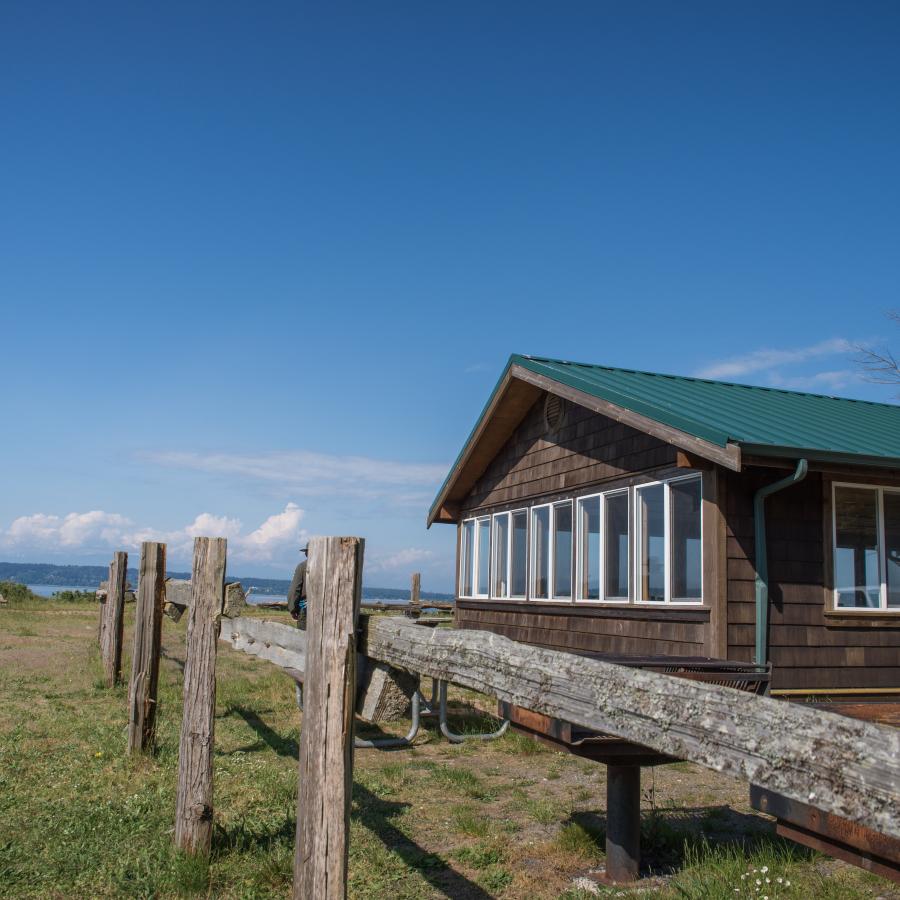 Picnic shelter at  Camano Island State Park. The shelter has a green, metal-looking roof, dark brown wooden siding, 5 white windows along the left side that take up the entire left wall, a single window visible on the entrance side of the building and a large, white framed door visible. The building is next to a picnic table, grill, and visible wooden post fence. In the far background you can just make out the Saratoga Pass and land on the opposite side of the water. 