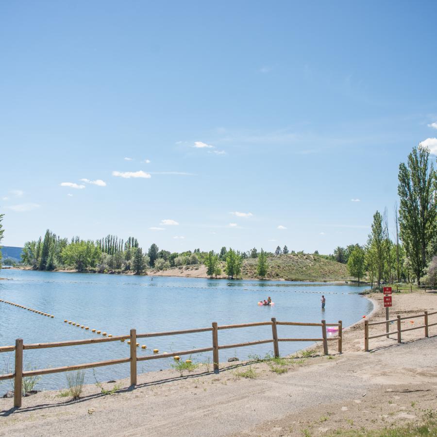 Buoy line in the water for the swim area and a sandy beach with a wooden fence lining the asphalt walkway