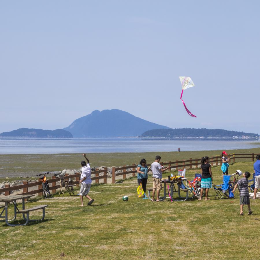 A group of kids and adults on a grassy lawn, lined with a wooden fence, prepare to fly kites in the wind. One kite is flying with a white cover and pink tail. The low tide water and islands are in the background.