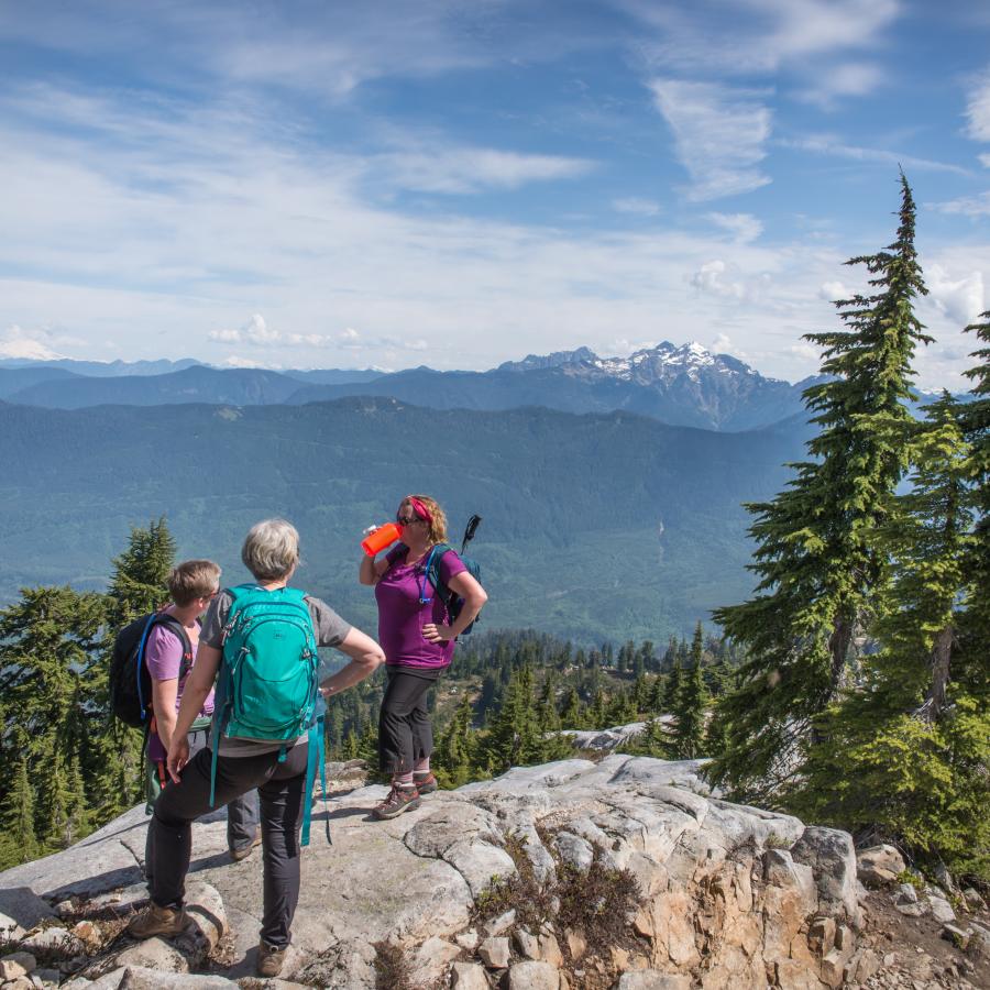 Hikers take a water break on a large boulder with evergreen trees scattered around them. Forested mountains and a snowcapped mountain peak sit in the background with a cloudy blue sky. 