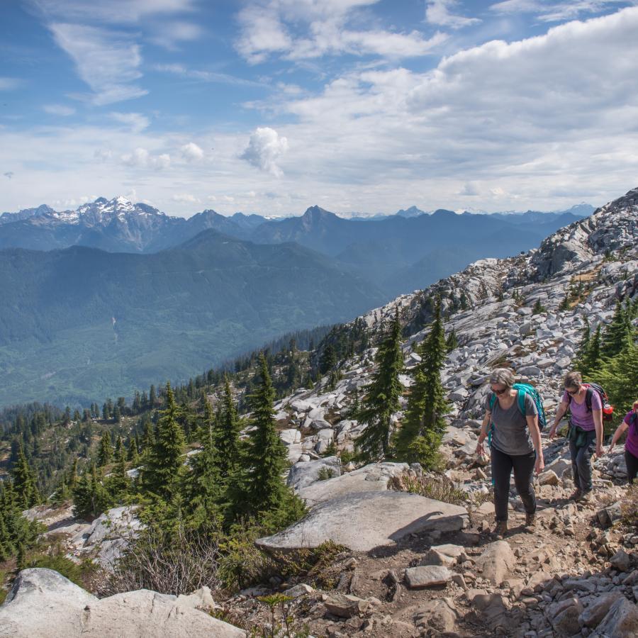 Three hikers hike the trail through dirt and rocks, surrounded by large boulders and evergreen trees scattered through the boulders. In the background is treed mountains with a large, snow capped mountain top with a blue cloudy sky overtop. 