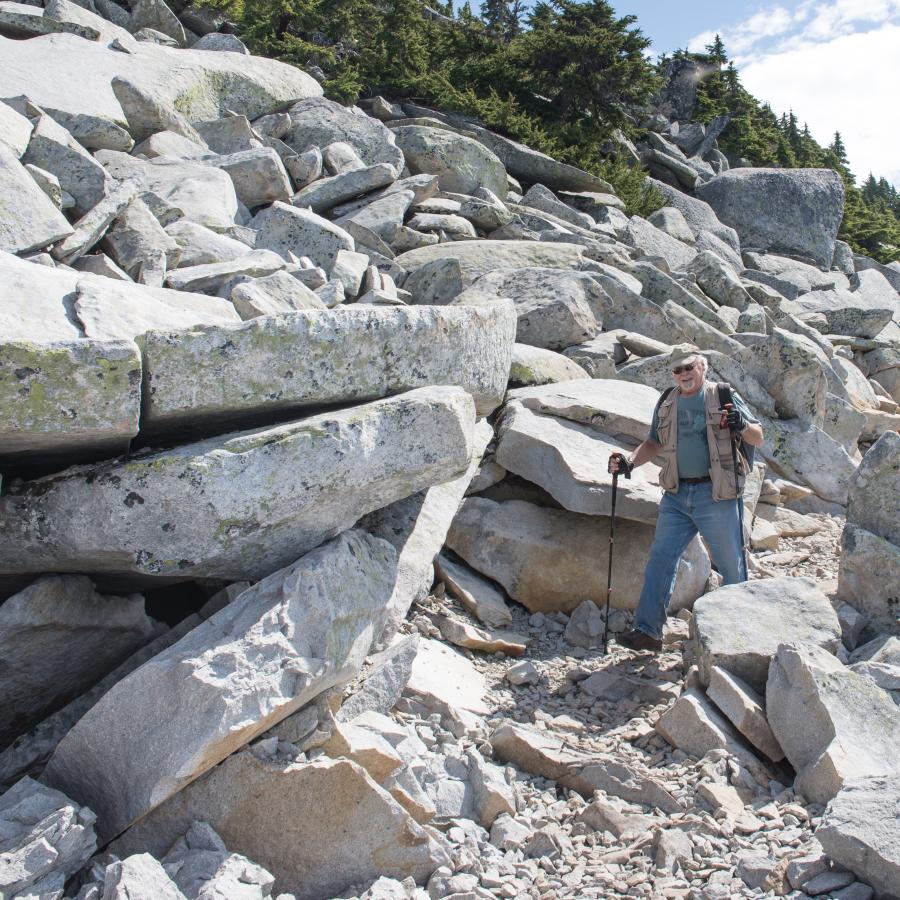 A smiling hiker coming down the rock covered trail, surrounded by large boulders and a few evergreen trees. 