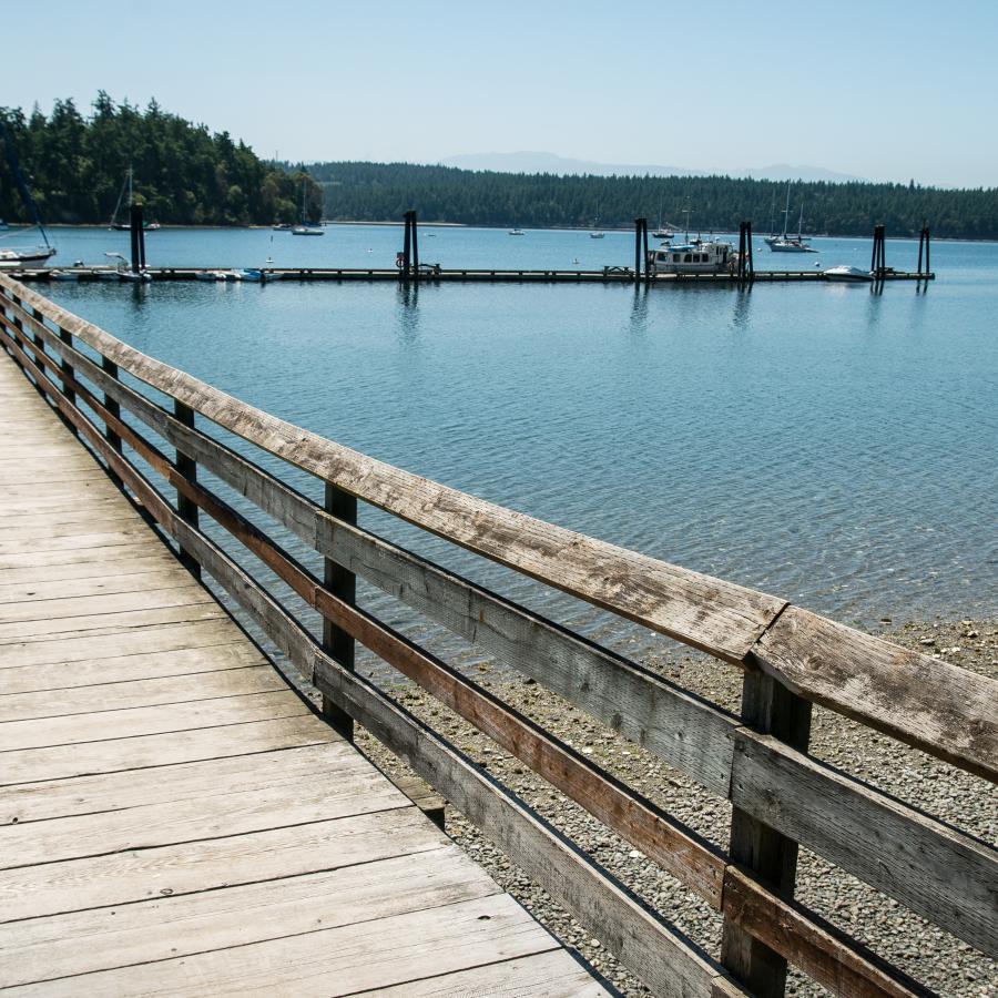 A T-shaped wooden dock sites in the water with boats tied to the end. In the background sits forested hills and a mountain poking out in the far back.