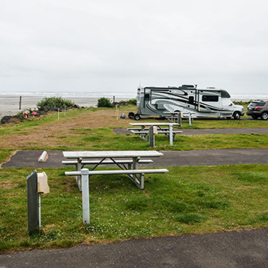 RV Camping parking spaces with picnic tables and small lawns right against the Pacific Ocean shore. A few RVs are parked in the spaces at the far end of the campground.