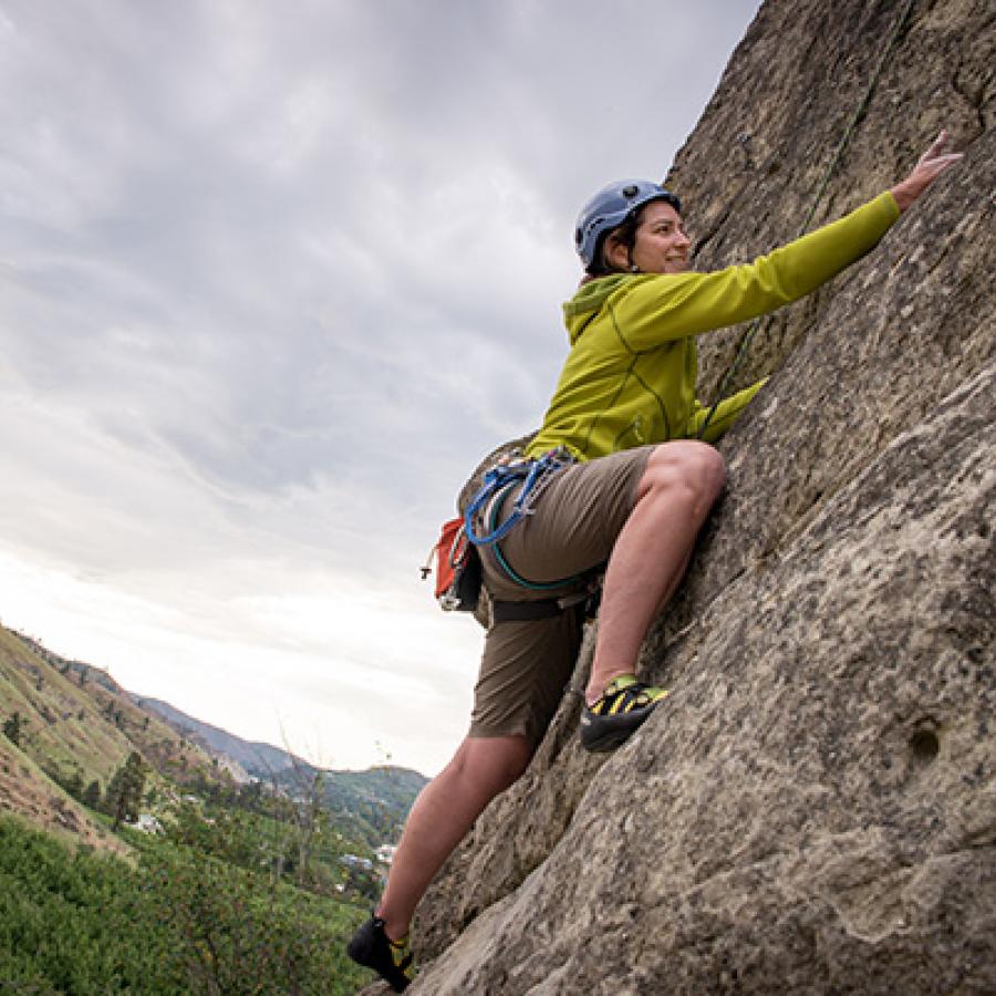 A person in a yellow shirt and shorts and wearing a helmet scales a granite rock face. A hillside rises in the distance under a cloudy gray sky.