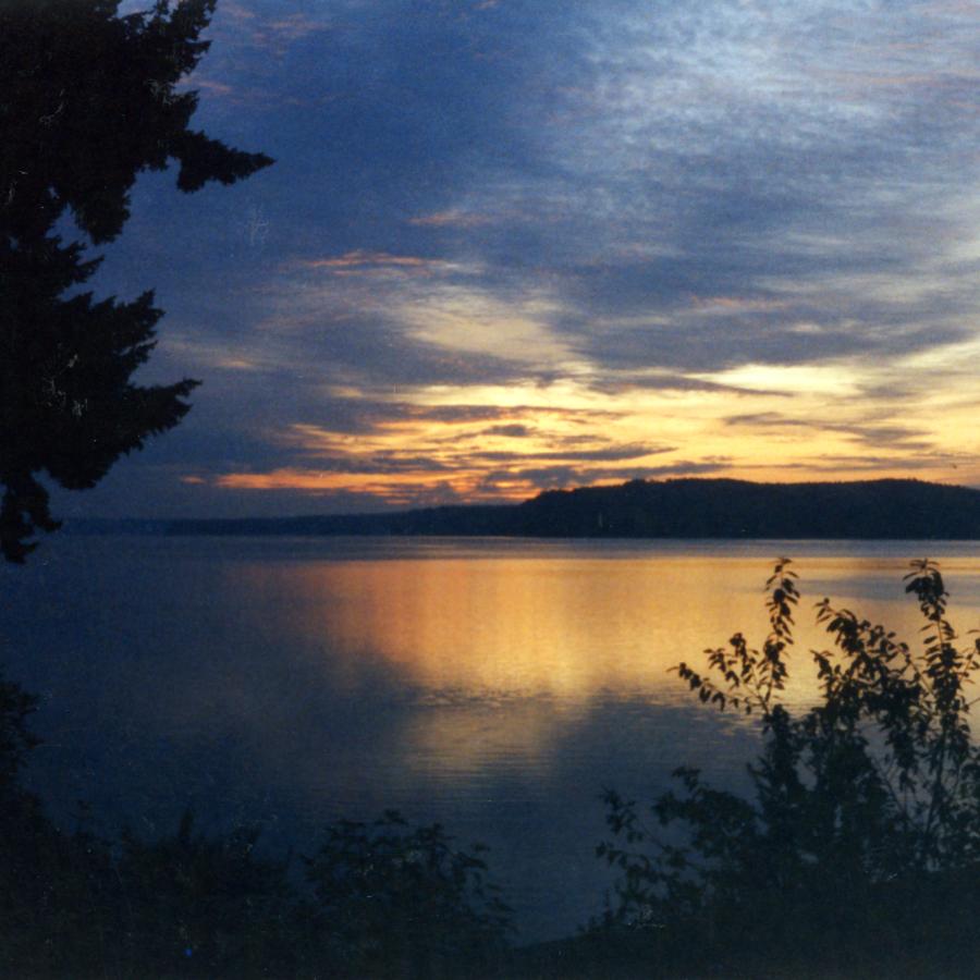 Dark shadows of trees and bushes frame an orange and dark gray late sunset reflected in the placid waters fronting Triton Cove State Park.