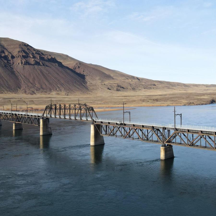 A n aerial shot of the Beverly Bridge, which spans the gray-blue waters of the Columbia River. A dark-tan, high-desert hilllside rises toward a pale blue sky in the background.