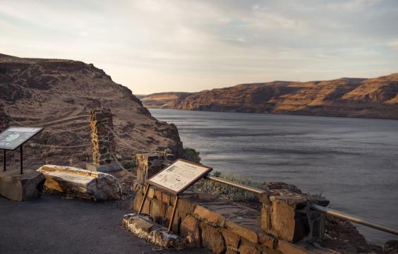 Interpretive panels sit in front of a rock wall with a chain rope with the Columbia River in the background.