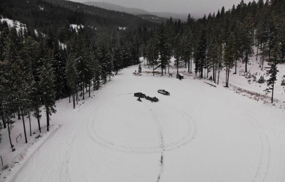 A open snow-covered area with circular tire tracks surrounded by trees with snowy hills in the background. A pit toilet stands in a corner.