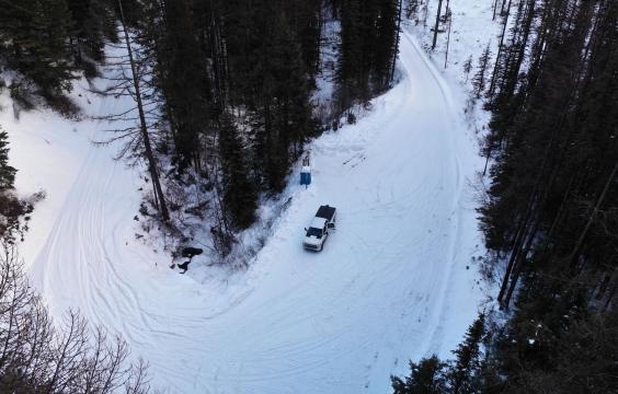 A forest road covered in thick snow and flanked by evergreen trees curves around a bend. A porta-potty and truck stand on the side of the road.