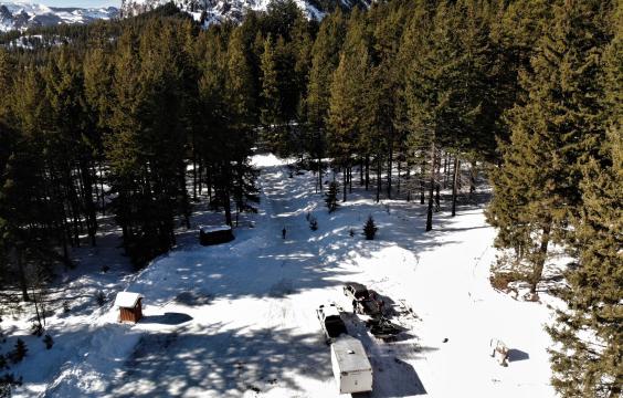 A truck with a trailer is parked next to a truck unloading two snowmobiles in a snow-covered parking area surrounded by pine trees. A porta-potty stands on the left side.