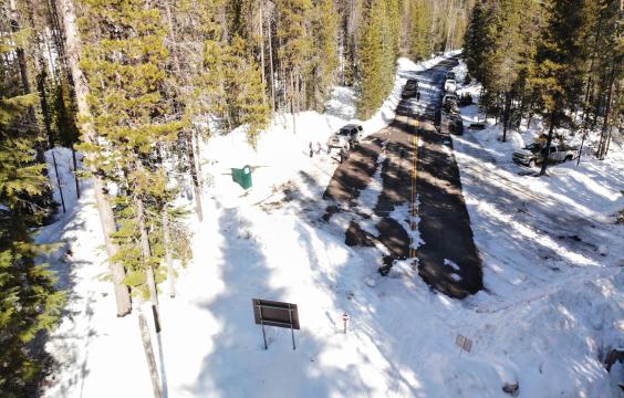 Plowed road with trees and cars parked on both sides ending at the edge of a snow-covered forest. A porta-potty lies on the left side.