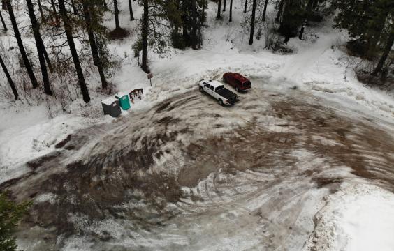 Overhead view of a plowed highway flanked by tall pine trees and snow-covered ground leading to a small plowed pull-off parking area.