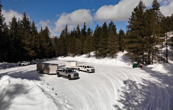 Two trucks with trailers sit in the middle of a plowed parking area covered by a layer of snow. A porta-potty stands on the right edge.