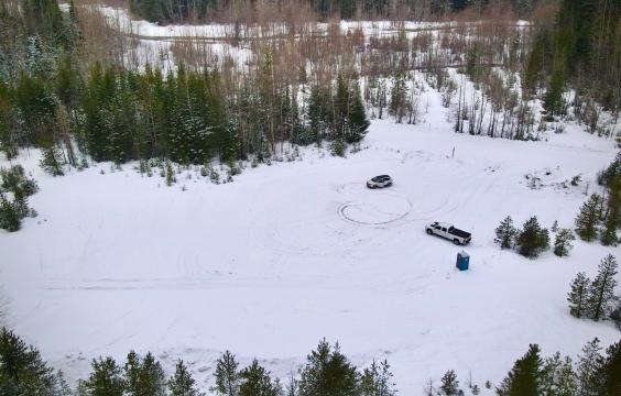 A large unplowed parking area bordered by young trees, with a narrow plowed road in the distance. A porta-potty is present.