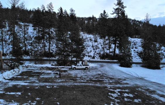 An icy forest road leads to a gravel parking lot with patchy snow next to a plowed forest road. A truck and porta-potty stand in the corner.