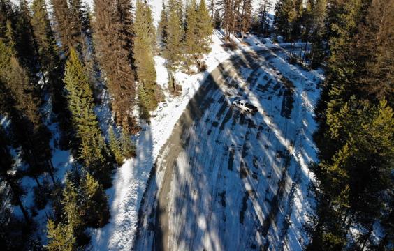 Overhead view of a plowed parking area covered by strips of thin snow and surrounded by pine trees.