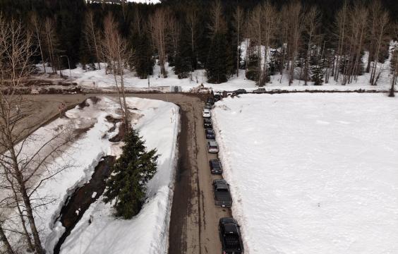 Paved forest road next to a highway, both plowed clean and bordered by thick unplowed snowy ground. A line of trucks and three porta-potties lie along the edge of the road.