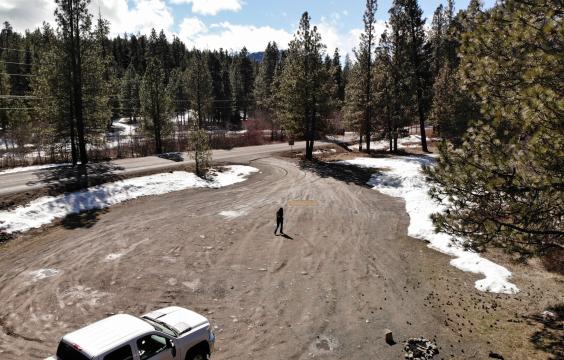 Dirt parking lot by the side of a highway surrouned by patchy snow and pine trees. A truck is parked in the foreground.