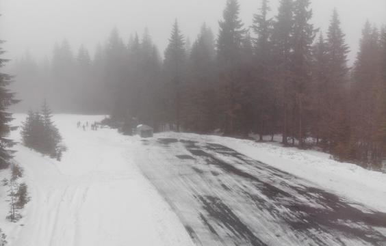 Foggy view of an icy parking area surrounded by thick snow-covered ground. A pit toilet hut and several skiiers stand on one end of the lot.