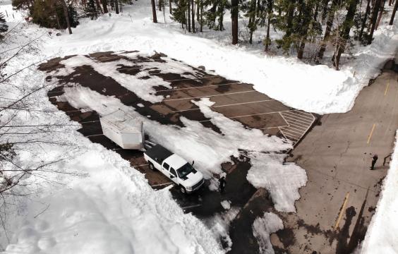 Paved parking area covered by patchy snow bordered by pine trees next to a plowed highway. The lot can fit a dozen trucks.