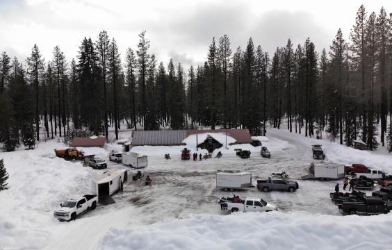 Snowy parking lot bordered by high piled up snow and pine trees. A long building half buried in snow lies on one side of the lot and a dozen trucks with trailers are parked along the perimeter.