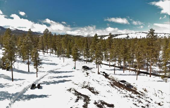 A snowy parking area with two snowmobiles and three trucks parked on the perimeter lies next to a plowed highway lined with pines.