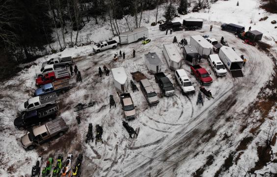 Trucks with trailers parked in the middle and along the edge of a parking lot covered by light snow surrouned by pine trees.