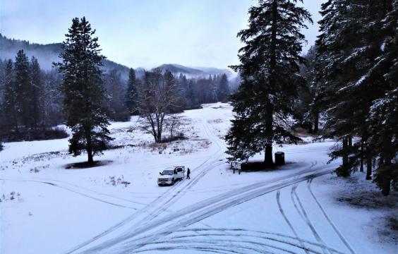 An intersection of two roads with thin snow, tire tracks lead away from the intersection on unmarked trails. A porta-potty sits under a tree, with misty mountains in the background.