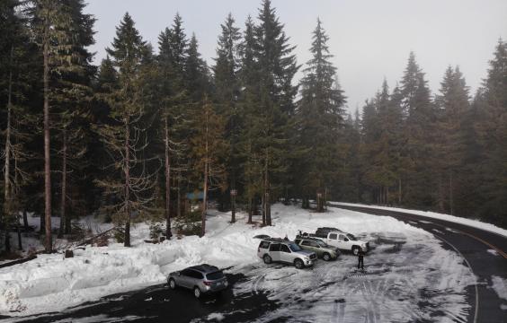 A plowed highway curves around a plowed parking area next to a forested trailhead.