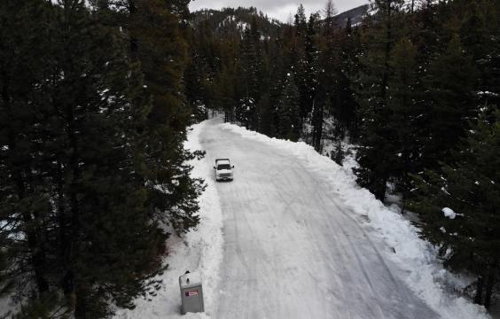 Plowed forest road covered in thin layer of snow with piled up snow and trees on both sides. A truck is parked on the left side.