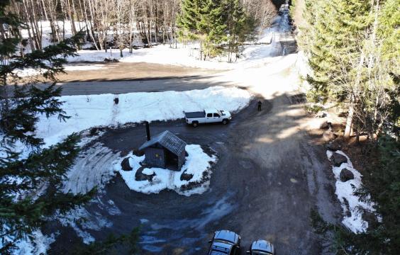 A bathroom hut in the middle of a plowed dirt lot surrounded by unplowed snow areas and trees.