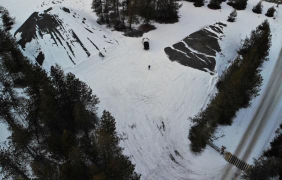 Parking area with compacted snow, flanked by two snow-covered pyramid-shaped hills and scattered trees along the edges. A plowed road appears in the corner.