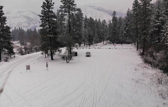 A snow covered parking lot surrounded by pine trees with snow-dusted mountains in the background.