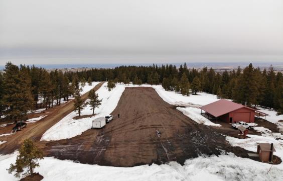 A long parking area next to a forest road, both plowed clean surrounded by snow-covered ground. A pit toilet hut sits in the corner and a small building with a garage and covered picnic area lie at the right edge.