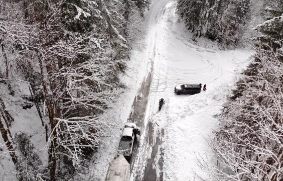 Thick snow covers a pull-off parking area along a forest road surrounded by a snowy forest. A truck with a trailer sits on the forest road and another truck sits on the pull-off.