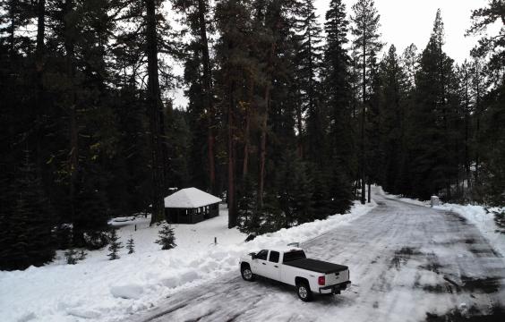 Icy forest road opening onto a wider ice-covered parking area bordered by unplowed snow and snowy ground. A picnic shelter sits a 50 feet back from the edge of the lot.