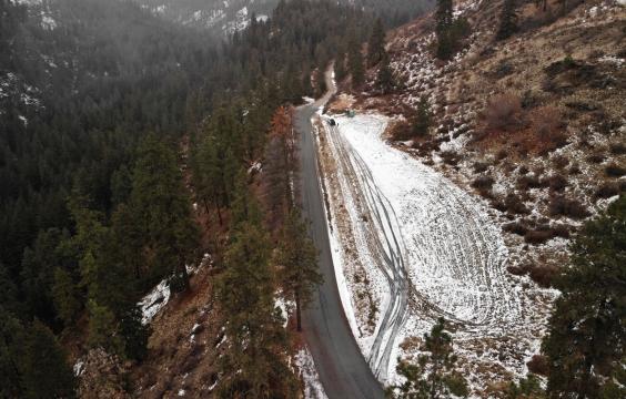 Overhead view of a long parking area covered by light snow running along a plowed highway, on a mountainside covered by pine trees. A porta-potty lies at one end.