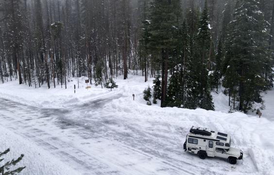 A forest road leading to a parking area, both covered in a thin layer of snow and ice, next to a snow-covered forest. 